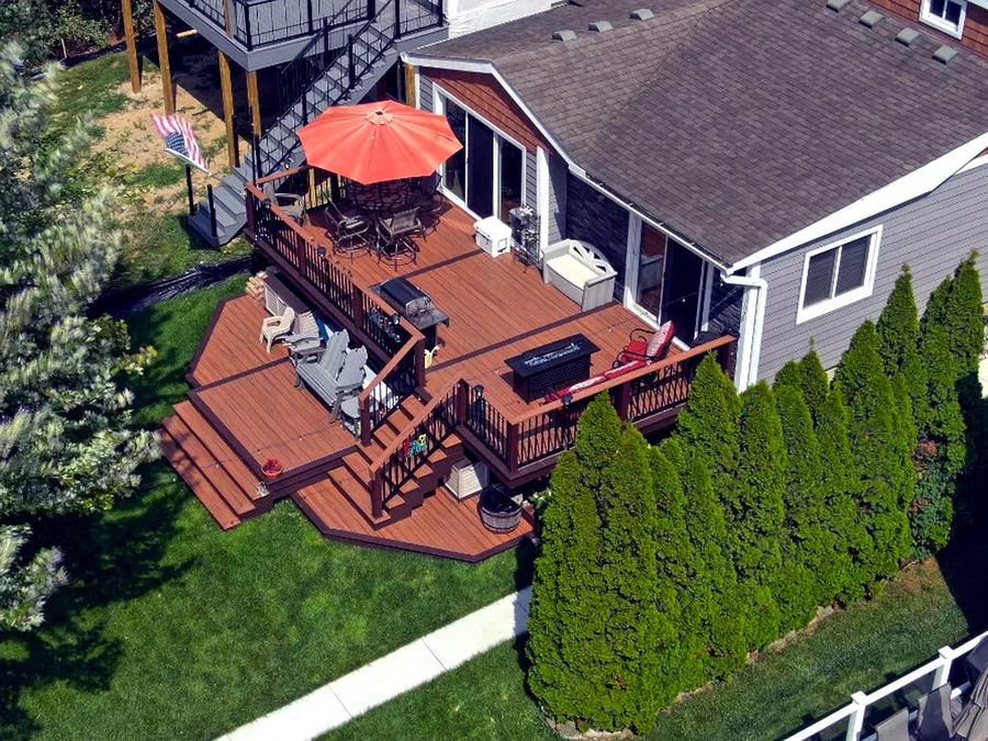 Drone aerial of a multi-level Trex composite deck with fire pit table and umbrella, overlooking a landscaped backyard